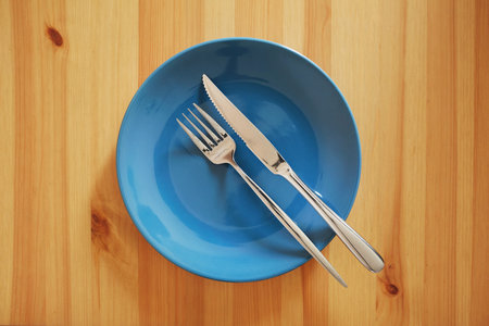 A signal "The meal was hearty and there was plenty of it". Empty and clean blue plate with fork and knife on a wooden table as an example of table etiquetteの写真素材