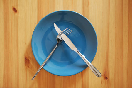 A signal "Meal break". Empty and clean blue plate with fork and knife on a wooden table as an example of table etiquetteの写真素材