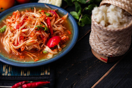 Spicy papaya salad served in a blue dish with sticky rice in a bucket and long beans, dried chilli and vegetables.の写真素材