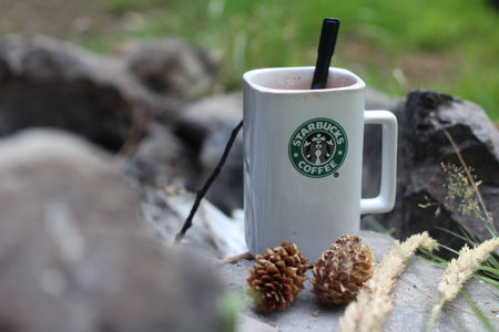 Washington, USA - January 13 2023 : Starbucks Coffee Mug Placed on rocks around a camping campfire with pine cones and reeds, modern tones.のeditorial素材