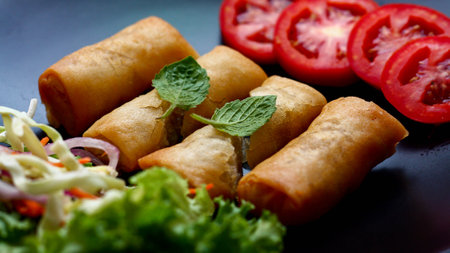 Fried spring rolls with vegetables and tomatoes placed in a black plate on a black wooden table and...