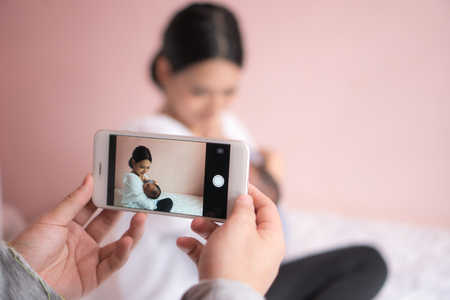 Hand taking pictures on a mobile phone of mother and baby. When the mother is lulling the baby. Pink background.の写真素材