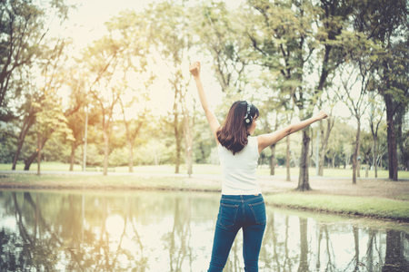 Beautiful girl is relaxing by listening to music. In the park there is a beautiful lake. She wears a white shirt. And she looks very happy. Vintage style. Copy space.の写真素材