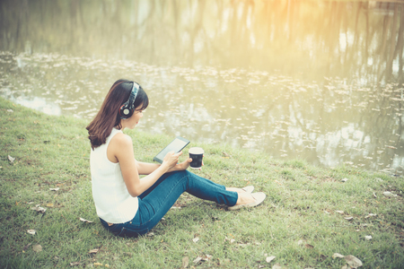 Beautiful girl is relaxing by listening to music. In the park there is a beautiful lake.She wears a white shirt And in her hands are choosing the song on the tablet. And she looks very happy.の写真素材