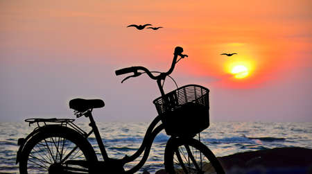Silhouette of bicycle and sunset sky with clouds Sun on water texture with ripples along seashore...