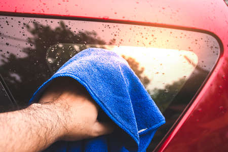 The human hand is wiping the water droplets on the car glass with a blue microfiber cloth