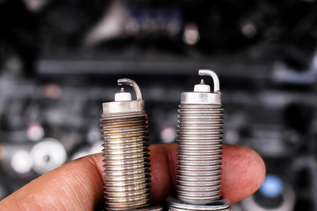 Comparison of used and new spark plugs, A hand holds old and new spark plugs in front of a blurred car engine. Automotive maintenance and ignition parts.の写真素材