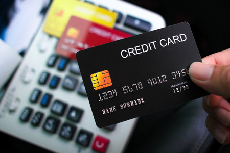 A hand holds a black credit card over a card reader or keypad, suggesting a transaction or payment...