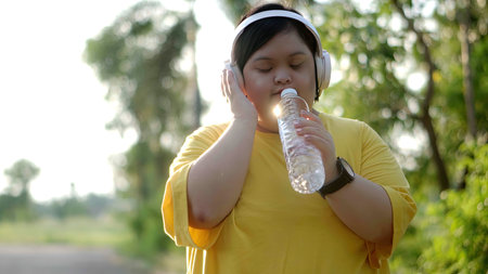 Asian girl with down syndrome in headphones uses water bottle as microphone to sing while exercising in the park.の写真素材
