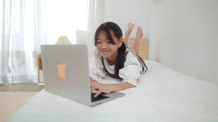 Happy little girl child. Cute little girl lie on his stomach a bed and watching something interesting on a laptop.の写真素材