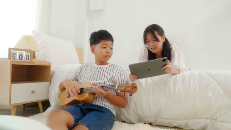 A girl lying in bed playing tablet and a boy sitting beside bed playing guitar in a bedroom.の写真素材