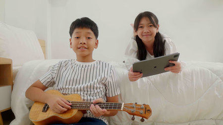 A girl lying in bed playing tablet and a boy sitting beside bed playing guitar in a bedroom.の写真素材