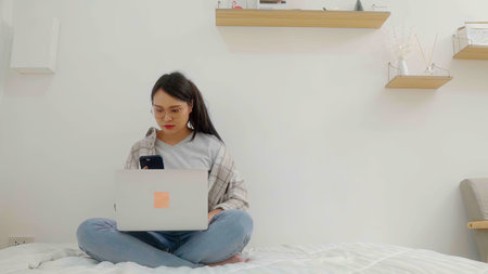 Asian  female wearing eyeglasses messaging with smartphone and a notebook laptop computer on bed at home.の写真素材