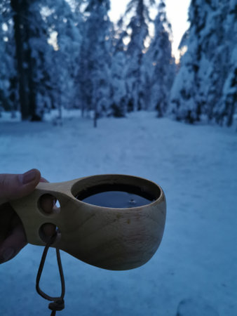 A kuksa wooden cup filled with hot tea on a fence with a snowy mountain view at morning sun.の写真素材