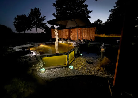 Night outdoor scene of a landscaped garden pond with illuminated water, decorative statue, glowing lanterns, gravel paths and wooden fence under deep blue evening sky, creating a calm and atmospheric ambianceの写真素材