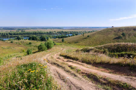 Country dirty road leads through green hills to a blue river