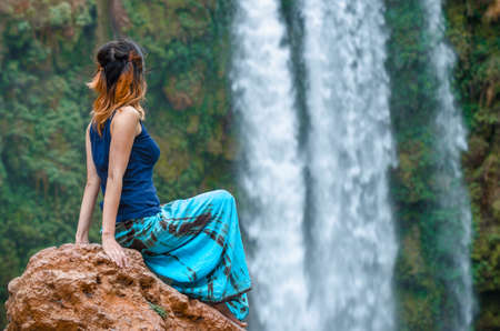 Girl sitting on rock and looking at waterfall background. Ouzoud falls in Morocco.の写真素材