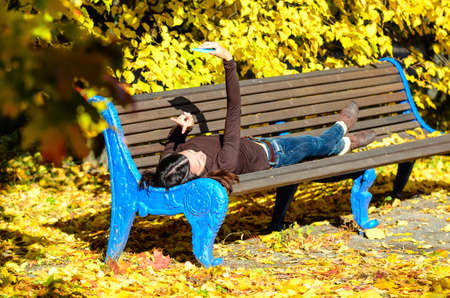 Attractive young woman laying on a brown wooden bench with mobile phone in her hands in a beautiful...
