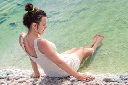 Sexy attractive girl in white dress sits near water of the Backunchak lake with her legs lowered into emerald sea. Young woman relaxing sitting on the edge of salt lake on white crystals of salt. Back to cameraの写真素材