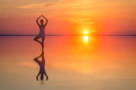 Beautiful female model open arms under the sunrise at seaside. Calm water of salt lake Elton reflects silhouette of woman. Sun goes under horizon. Girl stands alone in water. Sun reflects in Elton's water.の写真素材