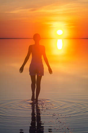 Beautiful female model open arms under the sunrise at seaside. Calm water of salt lake Elton reflects silhouette of woman. Sun goes under horizon. Girl stands alone in water. Sun reflects in Elton's water.の写真素材