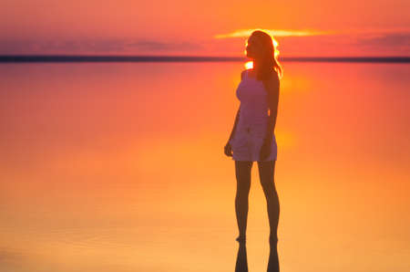 Beautiful female model stands in front of the sun under sunset at seaside. Calm water of salt lake Elton reflects silhouette of woman. Sun goes under horizon. Girl stands alone in water.の写真素材