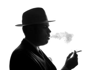 Silhouette of private detective lights cigarette. Agent looks like Al Capone stay side to camera. Police criminal scene in black and white. Gangster studio shotの写真素材