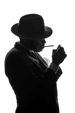 Silhouette of private detective lights cigarette. Agent looks like Al Capone stay side to camera. Police criminal scene in black and white. Gangster studio shotの写真素材