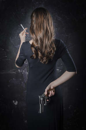 Noir film style woman in dress holding cigarette standing back to camera. Female posing with gun. Black and white photography. Old fashion photoの写真素材