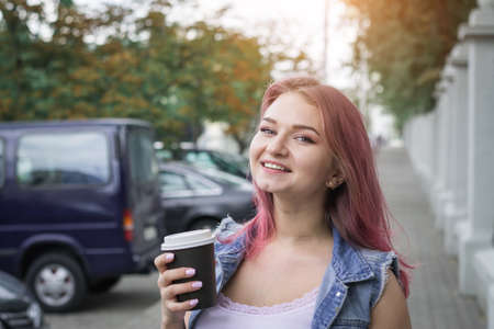 Portrait of a beautiful girl with red hair and a cup of coffee on a city street on a summer day