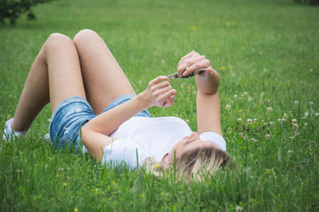 A girl lies on the grass and watches a message on her phone a summer day