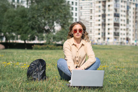 girl student sitting on the lawn on campus and working on a laptop on a sunny summer day