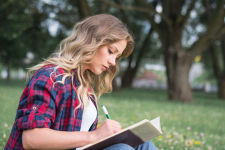 Beautiful, happy young student girl sitting on green grass near the backpack, bag under the tree...
