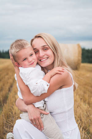 Young beautiful mother and son in white clothes posing for a photograph in a wheat field, happy family conceptの写真素材