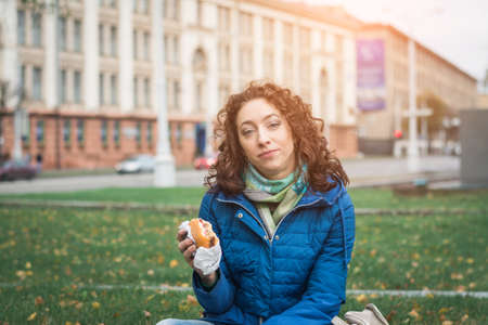 Girl student in a blue jacket eats a hamburger or cheeseburger on the street of her cityの写真素材