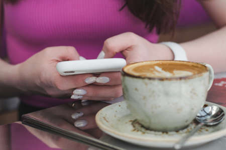 Young woman sitting in coffee shop at wooden table, drinking coffee and using smartphone