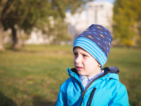Little Slavic boy child portrait in spring or autumn on the lawn in a city park in a hat and blue jacketの写真素材