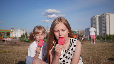 Two little girls sisters sit on the lawn in the city in the summer and eat delicious ice cream against the blue skyの写真素材