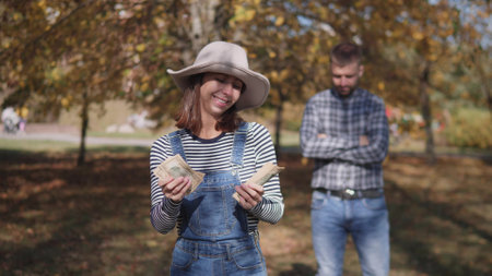 Joyful Woman Happily Holding Money in a Beautiful Autumn Park with a Pensive Manの写真素材