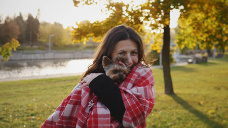 Woman Cuddles a Small Dog Wrapped in a Red Blanket During Sunset in a Park Surrounded by Trees and Waterの写真素材