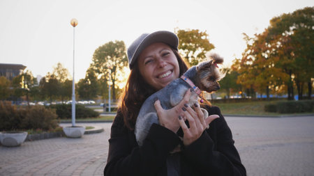 Woman Enjoys a Sunny Autumn Afternoon Holding a Small Dog in a Park Surrounded by Colorful Trees and Warm Golden Lightの写真素材