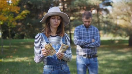 Woman Counts Cash in a Park While a Man Stands With a Concerned Expression During an Autumn Afternoon Filled With Colorful Leavesの写真素材