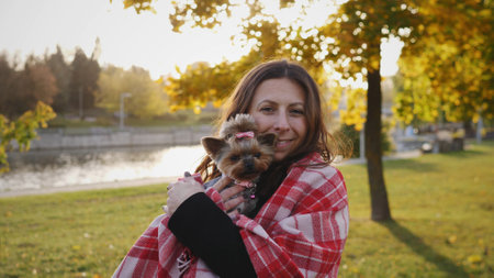 Woman Holding a Small Dog Wrapped in a Blanket While Enjoying a Sunny Autumn Day by the Lake in a Park Surrounded by Colorful Treesの写真素材