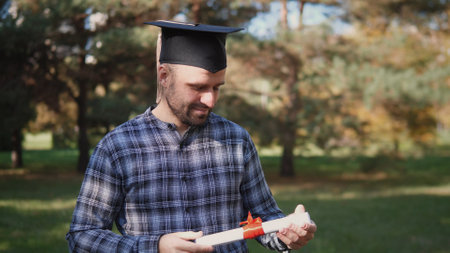 Graduate in a Cap Holding Diploma With Red Ribbon in a Park Surrounded by Trees During a Sunny Dayの写真素材