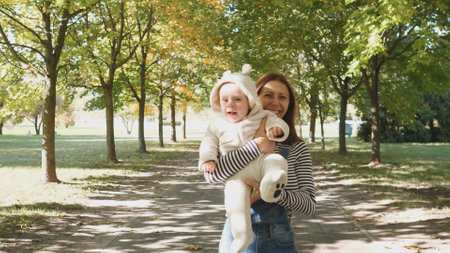 Mother Holds Baby in a Beautiful Park Surrounded by Colorful Trees on a Sunny Autumn Day While Sharing Joyful Moments Togetherの写真素材