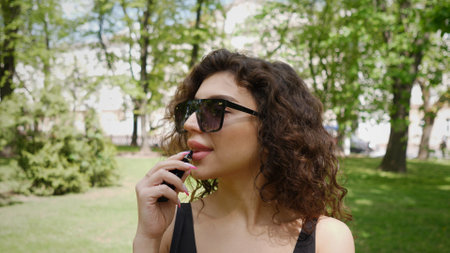 Young Woman Enjoying a Summer Day in a Park While Applying Lip Color in the Sunlight Surrounded by Greenery and Treesの写真素材