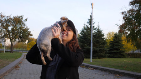 Woman Playfully Lifts Small Dog Under Clear Blue Sky in Peaceful Park During Autumn Afternoon With Colorful Leaves Surrounding Themの写真素材