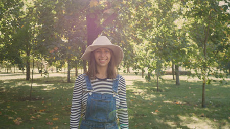 Woman Wearing Hat and Overalls Smiles in Sunlit Park Surrounded by Trees During Autumn Afternoonの写真素材