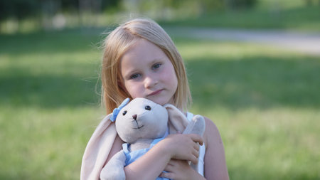 Little Girl Holding a Soft Bunny Toy in a Sunny Park During a Warm Afternoon, Enjoying Her Time Outdoors Surrounded by Greeneryの写真素材