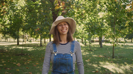 Smiling Woman in Striped Shirt and Denim Overalls Stands in Sunny Park Surrounded by Trees During Autumn Enjoying the Pleasant Weatherの写真素材
